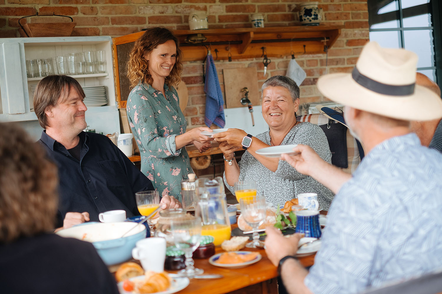 Eine lächelnde Frau verteilt Teller am Frühstückstisch für eine Gruppe von Freunden in der Sommerküche Domizil Kirschgarten in Metzelthin Uckermark